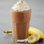 Thick dark chocolate protein shake topped with whipped cream and cocoa powder, served with sliced banana.