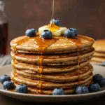 Stack of golden gluten-free pancakes topped with blueberries, maple syrup, and melting vegan butter.