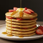 Stack of fluffy pancakes topped with fresh strawberries, melting butter, and syrup cascading down the sides.