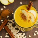 A golden smoothie in a glass mug with apple slices and a cinnamon stick on top, surrounded by oats and cinnamon powder.